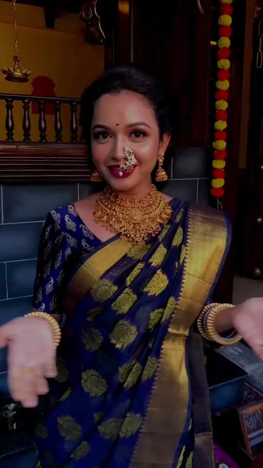 A portrait turn in a blue-and-gold Paithani beside the carved jharokha, marigold garlands hanging alongside.